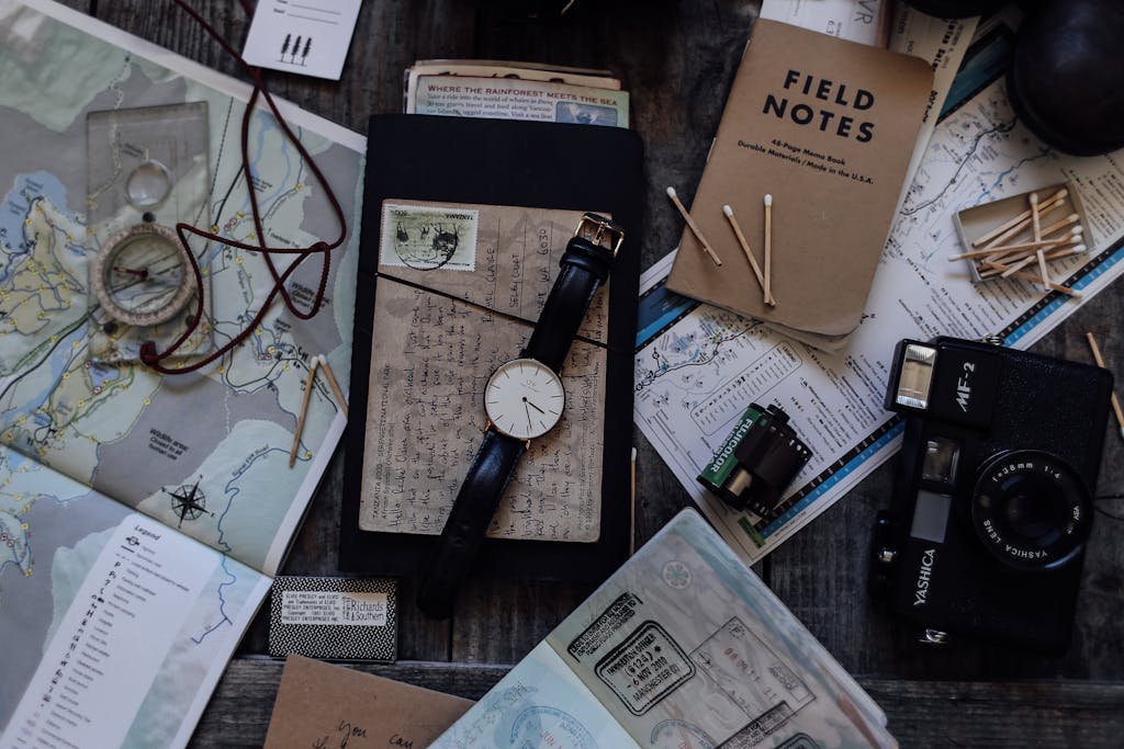 Flat lay of travel accessories including maps, vintage camera, and a wristwatch on a wooden table.